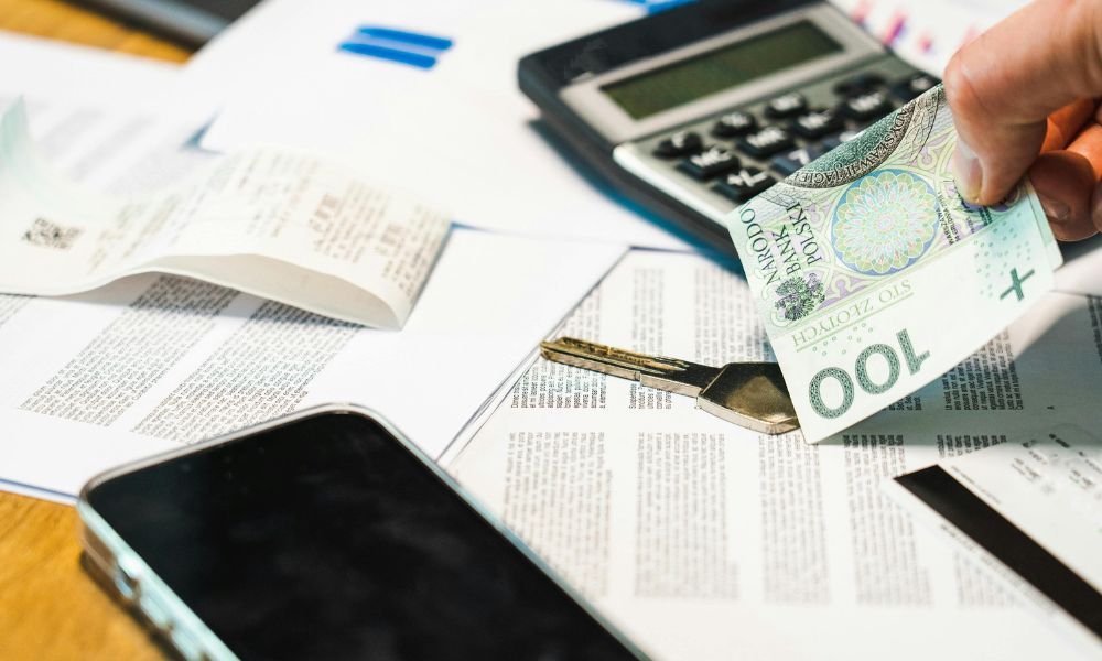 Hand holding a 100 zloty banknote above financial documents, receipts, a calculator, and a smartphone
