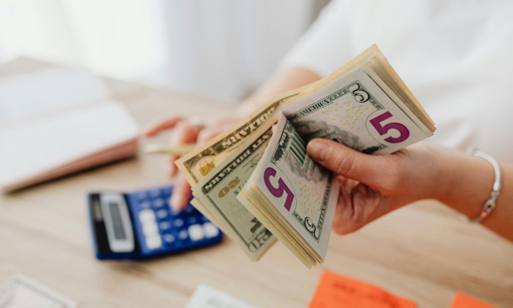 Person counting US dollar bills while using a calculator to review marketing and business expenses