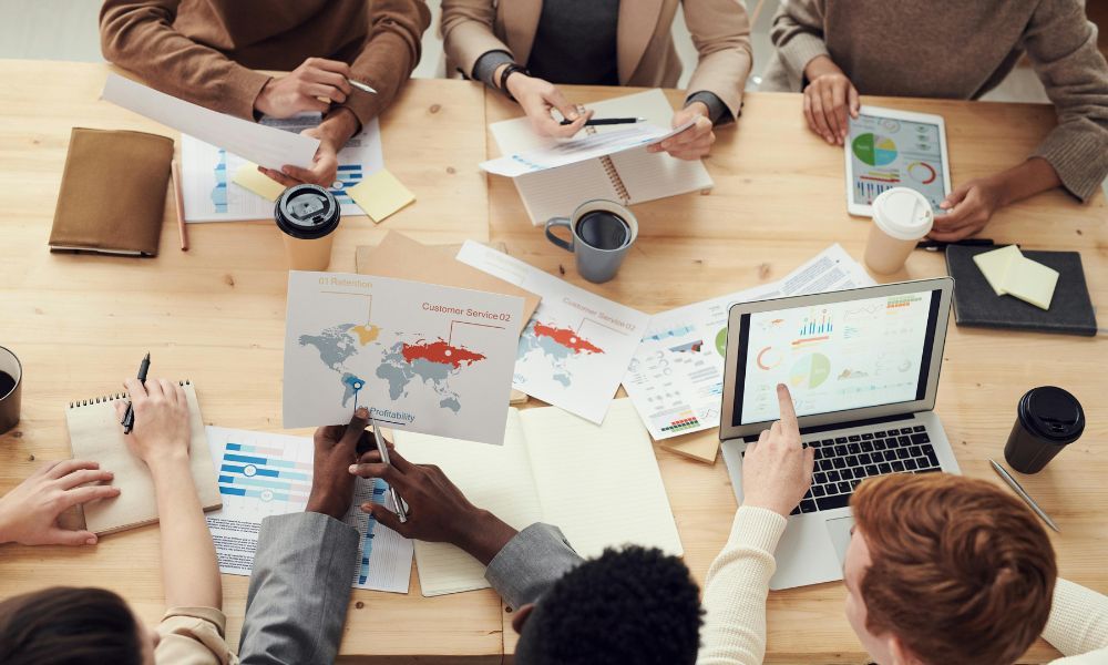 Top-down view of a team analyzing marketing reports, charts, world maps, and laptop dashboards spread across a wooden desk