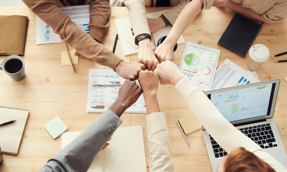 Team members joining their fists together above a desk with laptops, notebooks, and marketing reports with charts