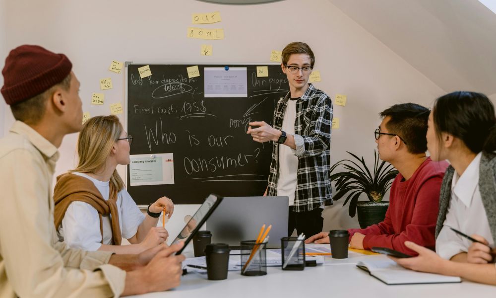 Startup team in a strategy meeting with a presenter pointing at a blackboard covered in sticky notes and questions like "Who is our consumer?"