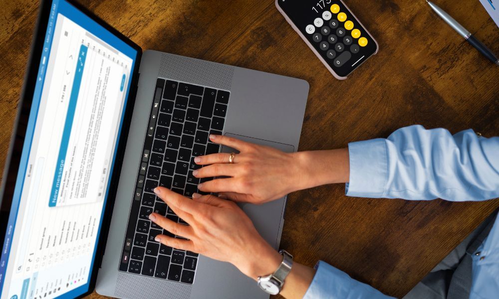 Top-down view of a person typing on a laptop with a new message draft on screen, a calculator on a smartphone, and a pen on a wooden desk