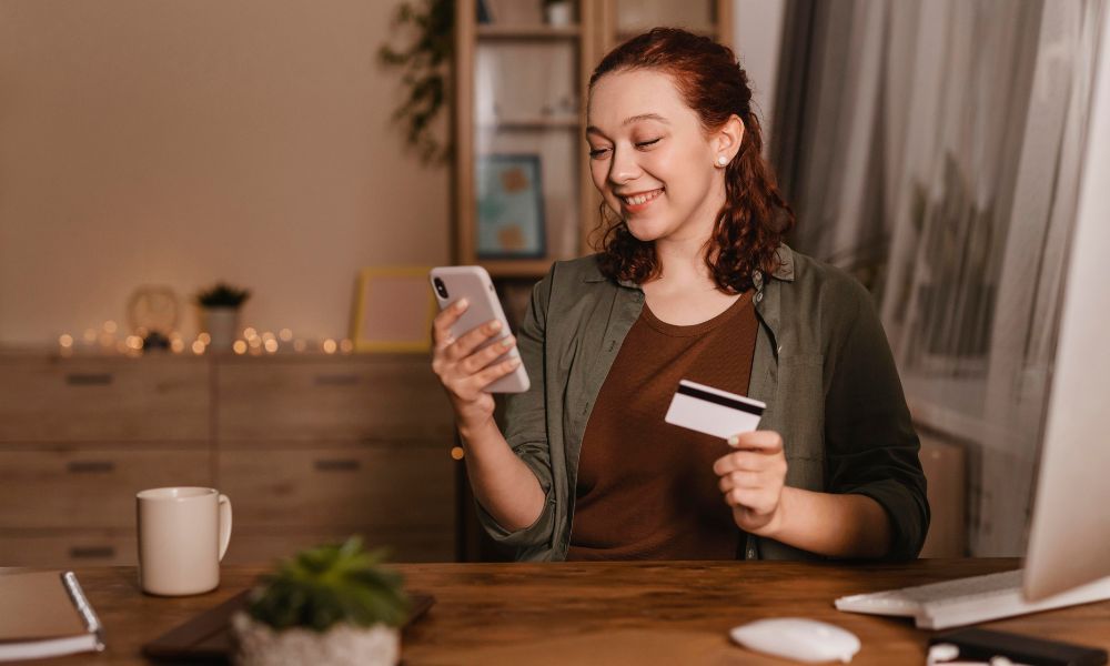 Young woman smiling at her phone while holding a credit card, sitting at a wooden desk with a coffee cup and computer monitor