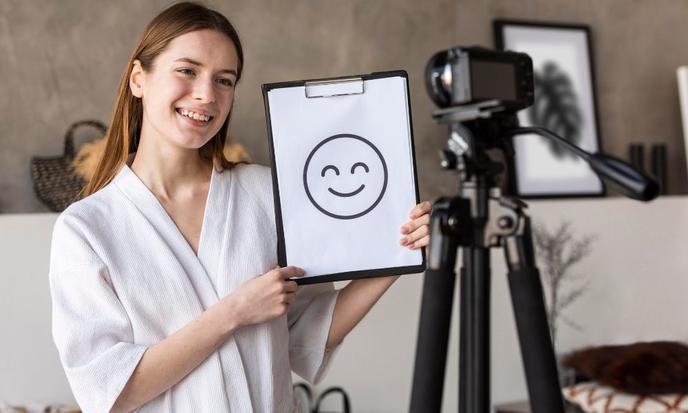 Young content creator holding a clipboard with a smiley face drawing in front of a camera mounted on a tripod