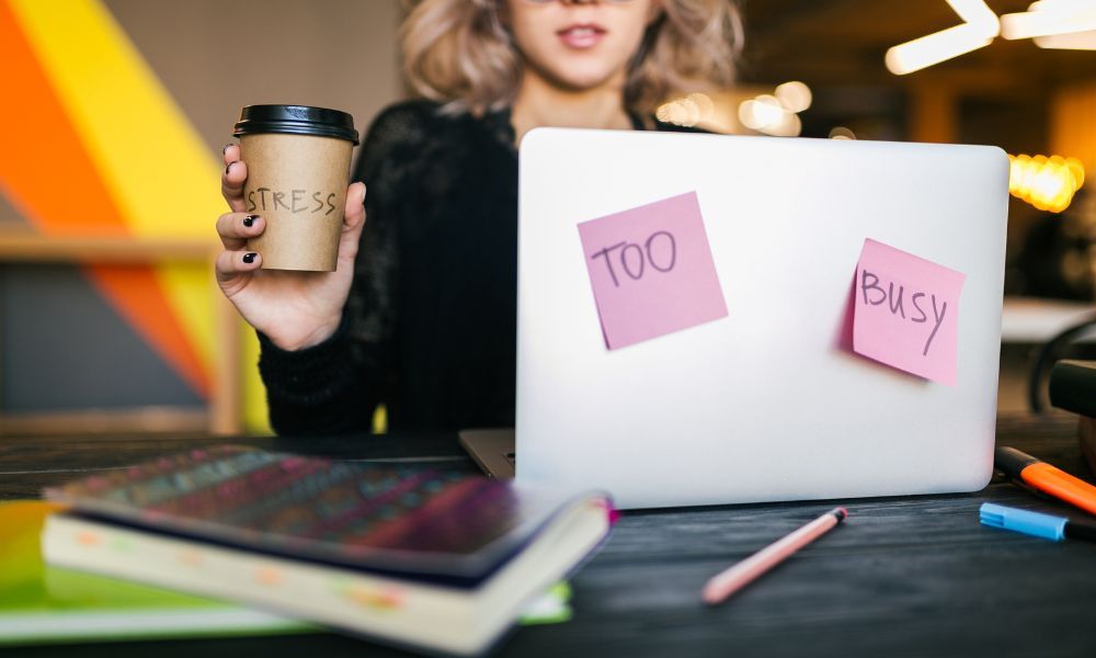 Stressed person holding a coffee cup labeled "STRESS" with sticky notes reading "TOO" and "BUSY" attached to a laptop